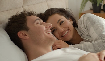 Casal sorrindo e se olhando com carinho deitados juntos na cama.