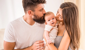 Pai e mãe beijando a cabeça de seu bebê recém-nascido que está no colo da mãe.